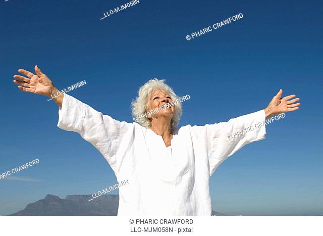 Senior woman looking up with arms outstretched on beach, Table Mountain in background, Table View, Cape Town, Western Cape Province, South Africa