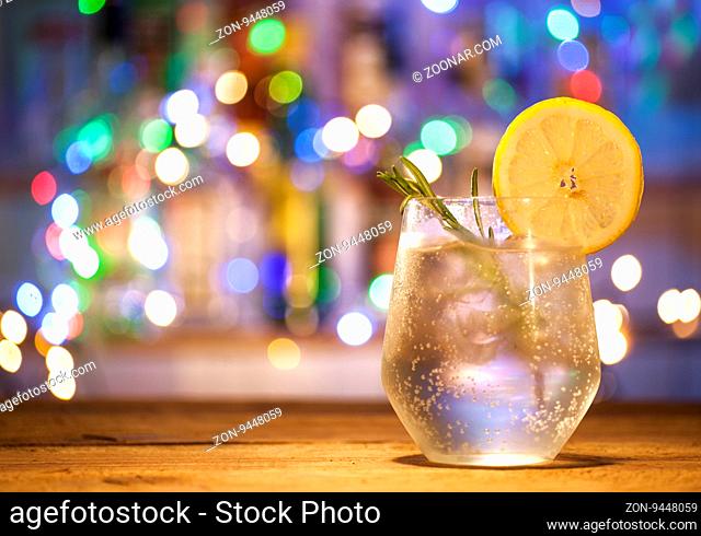 Glass with alcoholic drink with lime, rosemary and ice on wooden table and bar lights background