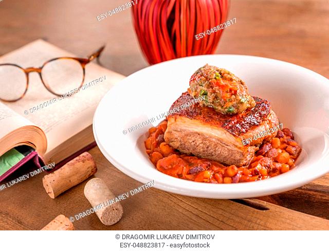 Pork belly with bean stew and mashed vegetables, served in a white plate, decorated with book and glasses on a wooden table