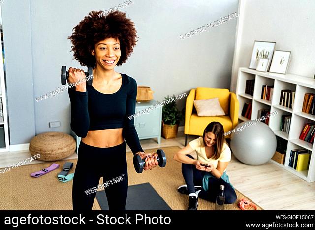 Happy young woman with dumbbell practicing by using mobile phone sitting in living room at home