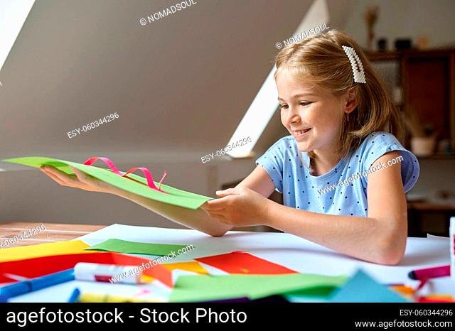 Little female child glues colored paper at the table, kid in workshop. Creativity lesson at the art school. Young painter, pleasant hobby, happy childhood