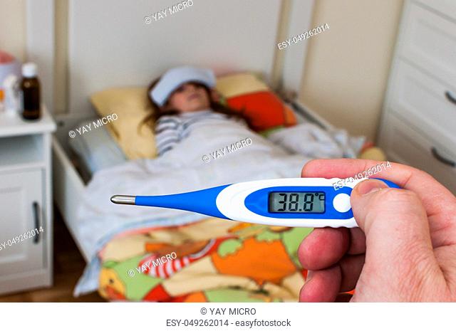 close up on a digital thermometer that marks 38 degrees Celsius. Little girl into bed with wet cloths on forehead