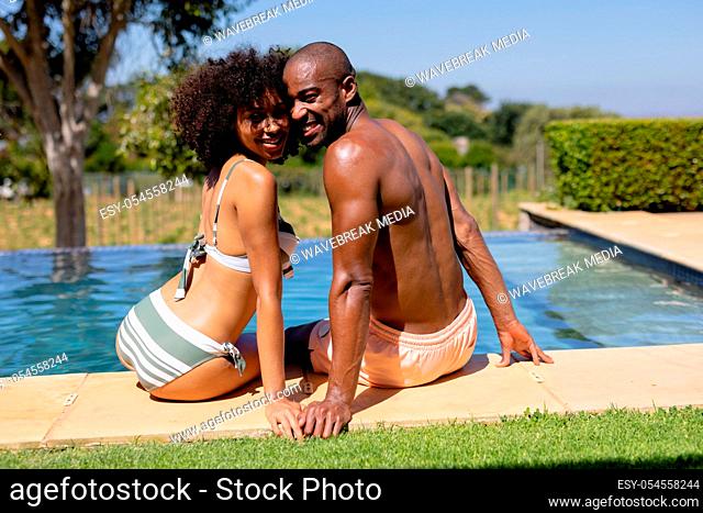 Rear view of a mixed race couple sitting in the garden on the side of a swimming pool wearing beachwear and sunbathing, turning around and smiling to camera