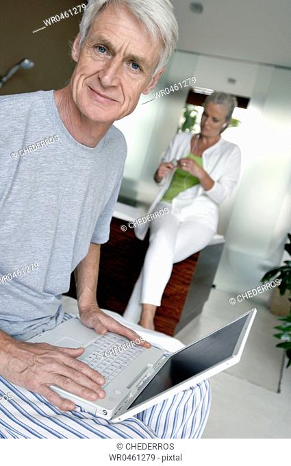 Portrait of a senior man using a laptop with a senior woman sitting in the background