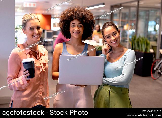 Portrait of happy biracial businesswomen standing with laptop and drink at modern workplace