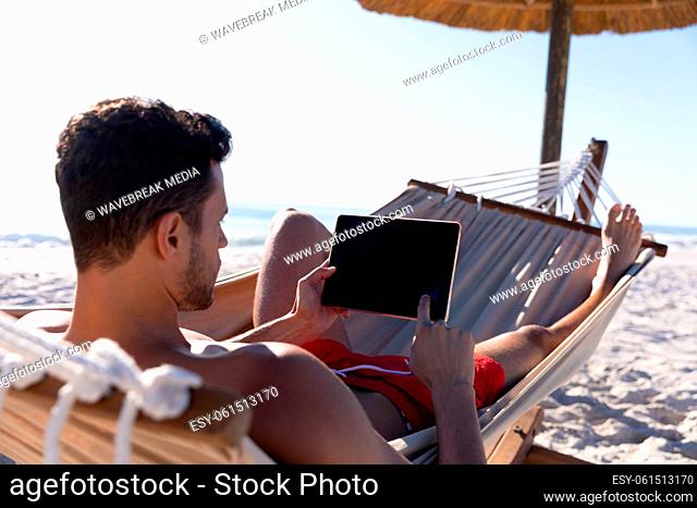Caucasian man lying on a hammock and using a digital tablet at the beach