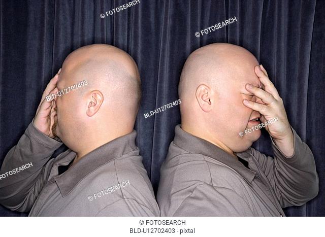 Profile of bald identical twin men standing back to back and grimacing with hands to head