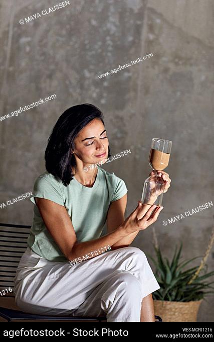 Businesswoman holding hourglass while sitting on chair against wall in office