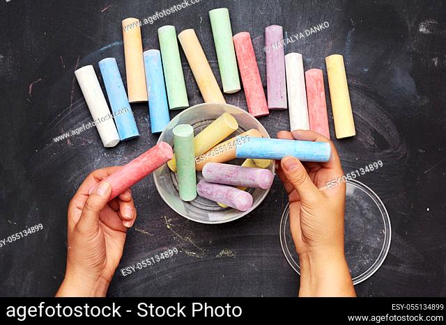 multicolored chalk in a plastic bucket on a black background, female hands fold objects into a container, top view