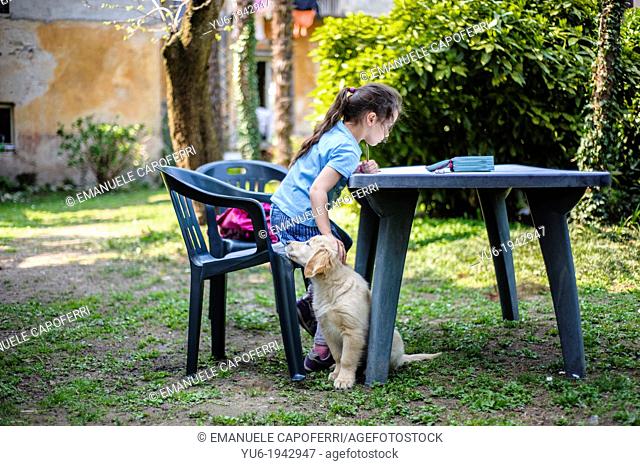 Little girl reads on the table in the garden while caressing a Golden Retriever puppy