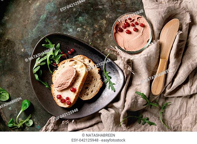 Chicken homemade liver paste or pate in glass jar with sliced whole grain bread, wood knife, cranberries, green salad served on ceramic plate with textile over...