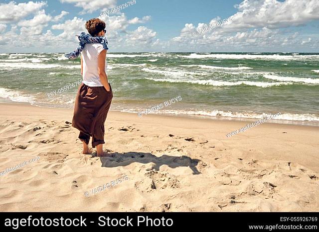 Middle aged woman standing on the beach looking at waving sea on windy summer day