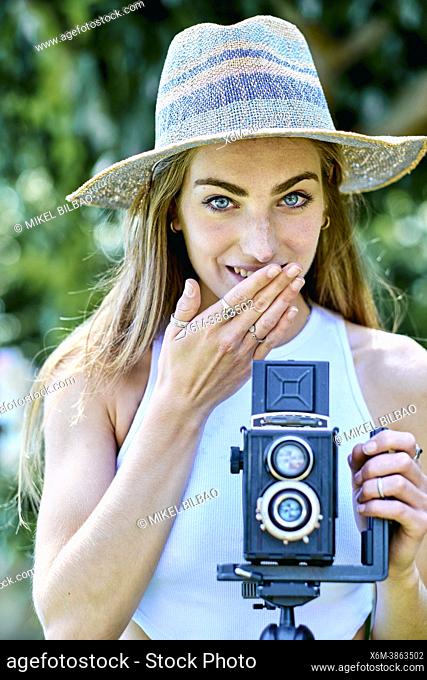 Portrait of a young beautiful caucasian woman in her 20's with a gesture of shyness wearing a hat and photographing with an old vintage camera on a tripod...