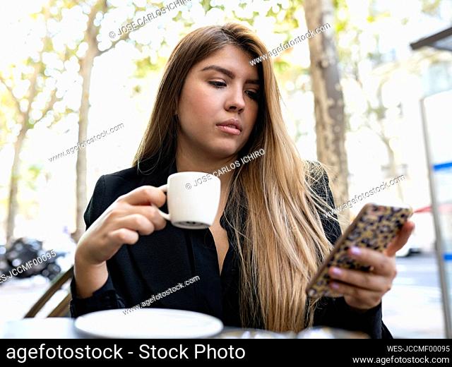 Woman using mobile phone while drinking coffee at sidewalk cafe in city