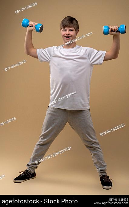 Full-length portrait of a cheerful teenager with a pair of dumbbells posing for the camera against the beige background