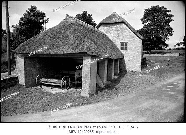 Exterior view of a single-storey thatched barn attached to a stone building with thatched roof and housing a wooden vehicle, likely a cart or trap
