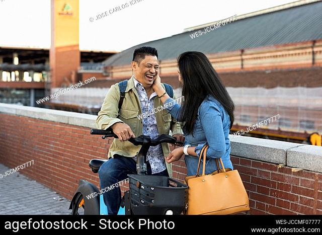 Woman touching face of happy man sitting on electric bicycle by wall
