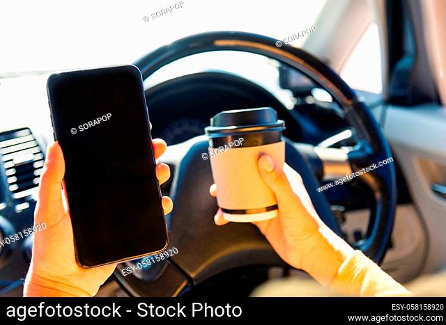 Asian woman drinking hot coffee takeaway cup inside a car and using smartphone blank screen while driving the car in the morning during going to work on highway