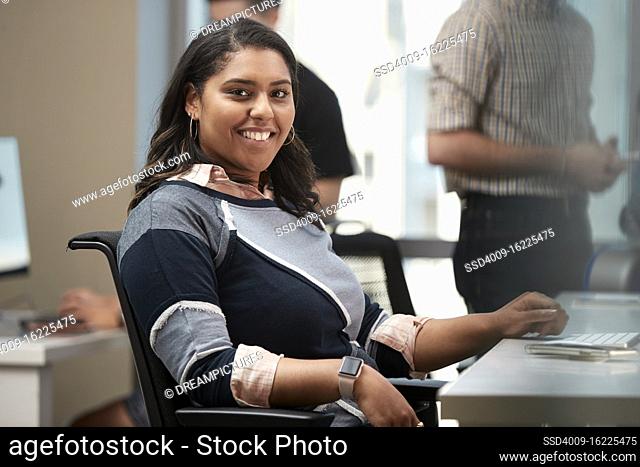 Young woman sitting at computer in office looking to the camera smiling, co-workers having discussion around dry erase board in background