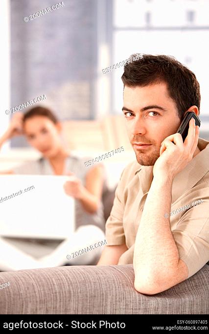 Young man sitting on sofa at home, talking on mobile, woman using laptop in the background