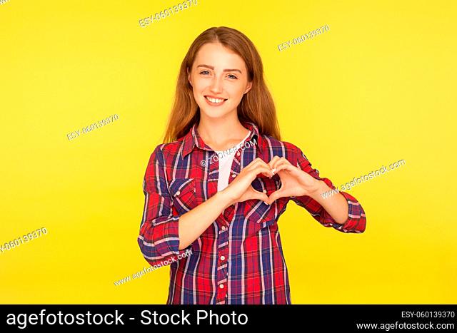 Portrait of beautiful lovely ginger girl with kind smile and in checkered shirt showing heart shape with hands, gesturing love, charity and hope concept