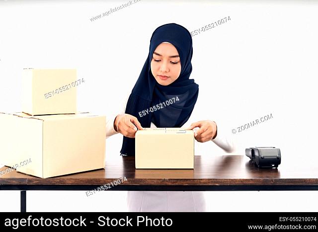 Asian muslim women merchant working with laptop computer from home on wooden table with postal parcel cardboard boxes preparing for shipping her order