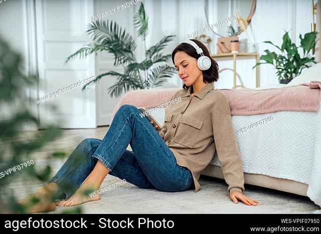 Smiling woman wearing wireless headphones listening to music and using mobile phone at home