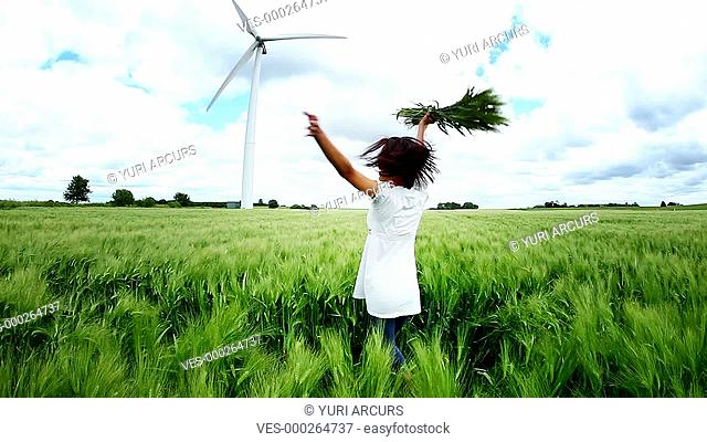 Young woman standing and twirling in a wheat field with a windmill behind her while she smiles