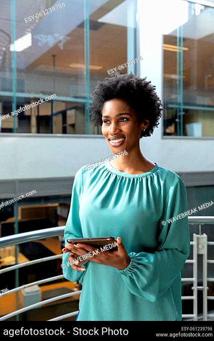 Smiling african american businesswoman holding tablet pc looking away standing at modern workplace