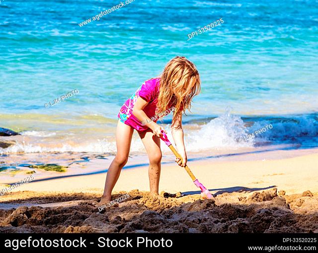 A young girl stands at the water's edge on Kapalua Beach on Kapalua Bay digging in the sand with a shovel; Kapalua, Maui, Hawaii, United States of America