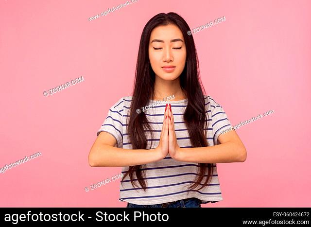 Calmness, harmony and yoga practice. Portrait of peaceful girl with long brunette hair in striped t-shirt holding arms in namaste gesture and meditating
