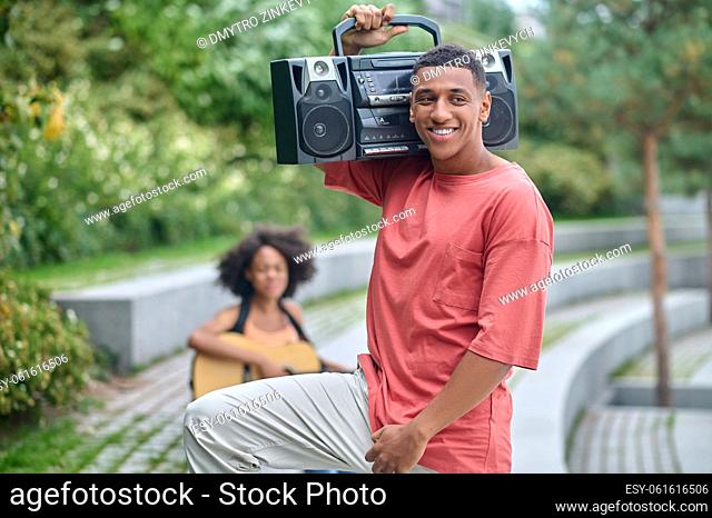 Enjoyment. Attractive dark-skinned guy with record-player on shoulder standing smiling to side and girl sitting behind in park