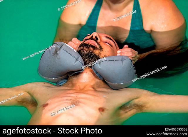 Female therapist giving facial water massage to man with closed eyes relaxing with rubber pillow around his neck in swimming pool