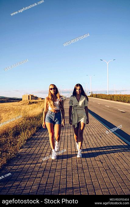 Young female friends walking on sidewalk against sky on sunny day