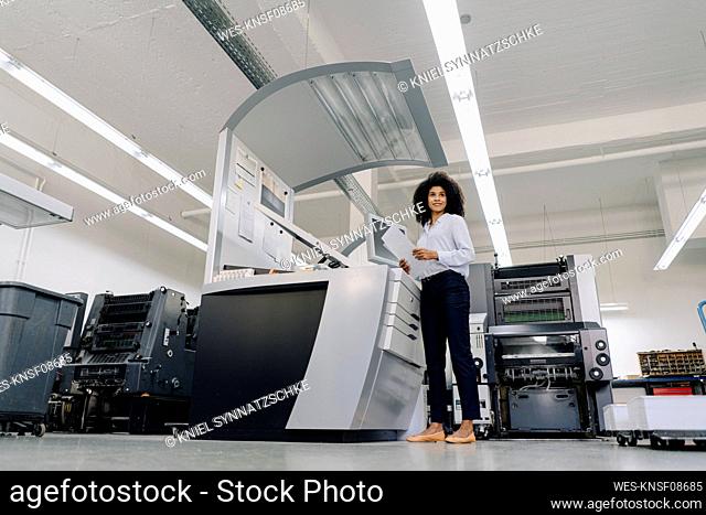 Smiling businesswoman with document standing by machinery in industry