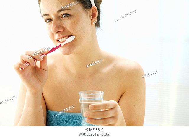 young woman dressed with a towel is cleaning her teeth with a toothbrush and is holding a glass of water in her hand