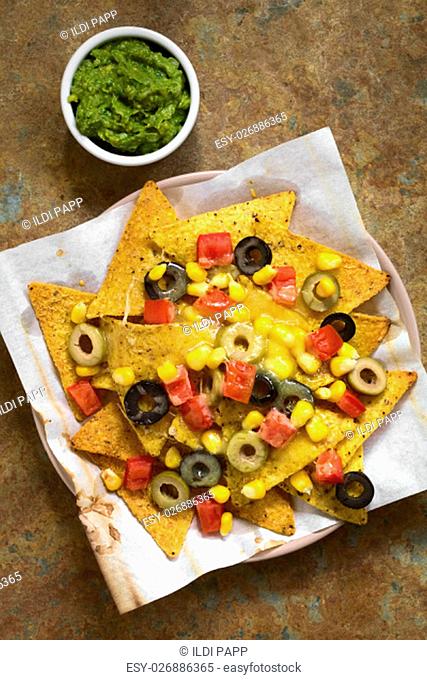 Baked nachos with cheese, green and black olives, red bell pepper and corn, with guacamole on the side, photographed overhead on slate with natural light