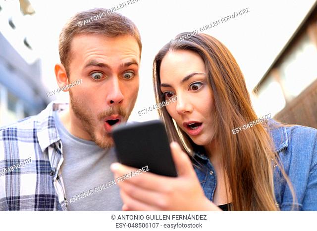 Front view portrait of surprised couple checking online news on smart phone in the street