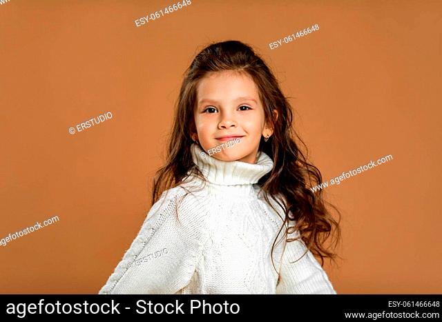Cute smiling little child girl in white sweater looking to camera on beige background. Human emotions and facial expression