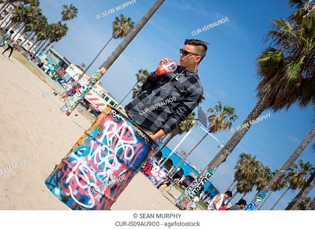Angled view of teenage boy drinking smoothie next to graffiti covered rubbish bin, Los Angeles, California, USA