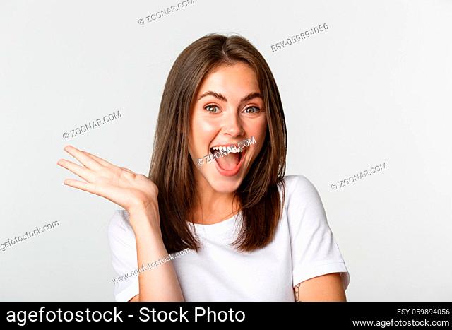 Close-up of excited beautiful brunette girl looking amused, waving hand to say hello, white background