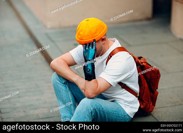 Young disabled upset sad man with artificial prosthetic hand sitting on sidewalk city street outdoor