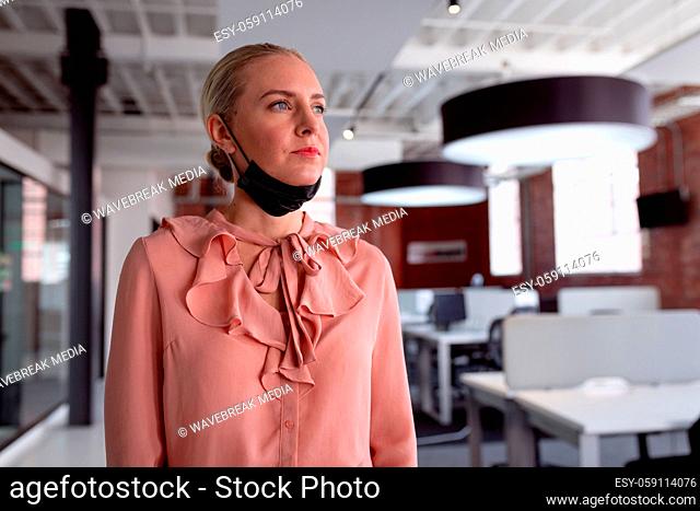 Thoughtful caucasian businesswoman wearing lowered face mask standing in office looking away
