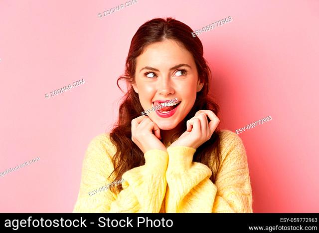Young devious girl licking her lips and teeth, looking aside with thoughtful face, having interesting idea, thinking, standing against pink background