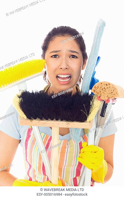 Distressed young woman in apron and rubber gloves holding cleaning tools