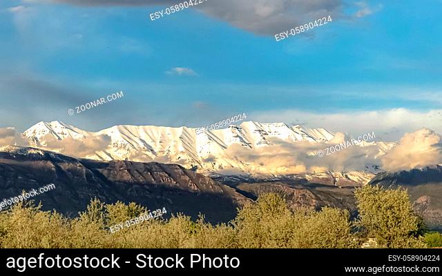 Panorama frame Scenic nature view with majestic snow peaked mountain under cloudy blue sky. Trees with lush green leaves can be seen in foreground on this sunny...