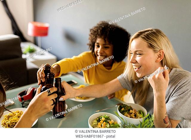 Happy girlfriends sitting at dining table clinking beer bottles
