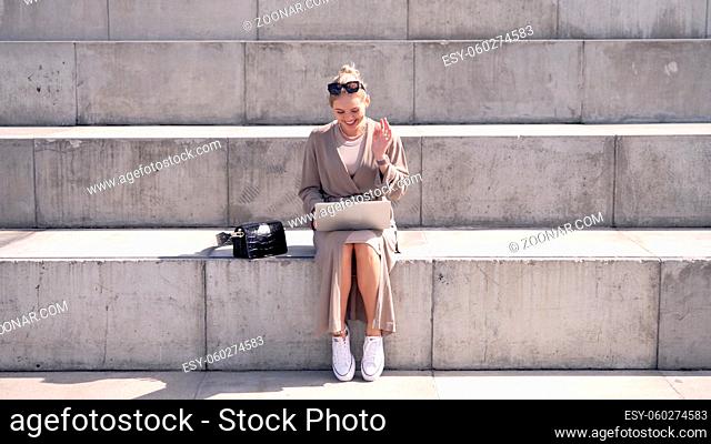 Stylish young female with laptop smiling and waving hand while sitting on stairs and making video call on sunny day on street