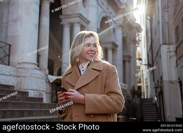Waist-up portrait of a smiling beautiful blonde with the smartphone standing among old architectural buildings in the city center