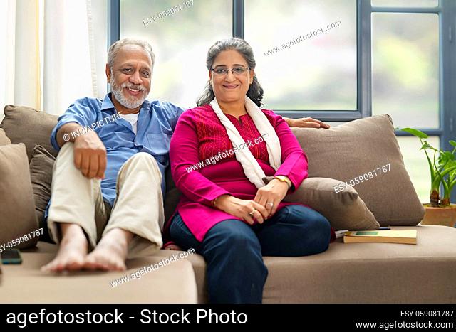A HAPPY OLD COUPLE SITTING TOGETHER ON SOFA AND LOOKING AT CAMERA